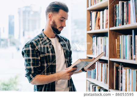 Young male librarian in casual clothing making order near bookshelves in college campus 86053595