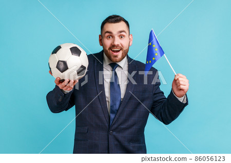 Excited man wearing official style suit, holding black and white soccer ball and flag of EU, watching football and support favourite team. Indoor studio shot isolated on blue background. 86056123