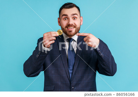 Attention to cryptocurrency. Satisfied businessman wearing official style suit pointing bitcoin and smiling to camera, showing digital money. Indoor studio shot isolated on blue background. Attention to cryptocurrency. Satisfied businessman wearing official style suit pointing bitcoin and smiling to camera, showing digital money. Indoor studio shot isolated on blue background. 86056166