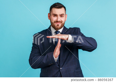 I need more time. Portrait of frustrated brunette man with beard in dark suit showing time out gesture, looking at camera, hurry to meet deadline. Indoor studio shot isolated on blue background. I need more time. Portrait of frustrated brunette man with beard in dark suit showing time out gesture, looking at camera, hurry to meet deadline. Indoor studio shot isolated on blue background. 86056167