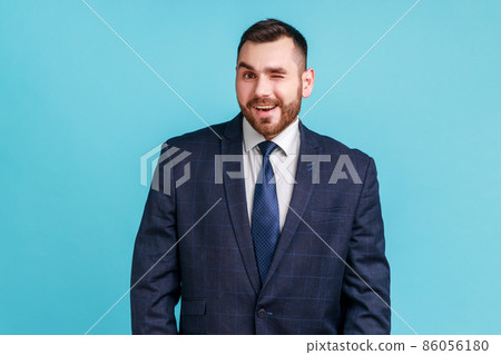 Optimistic young adult man with beard wearing official style suit standing looking at camera and winking, expressing positive emotions. Indoor studio shot isolated on blue background. 86056180