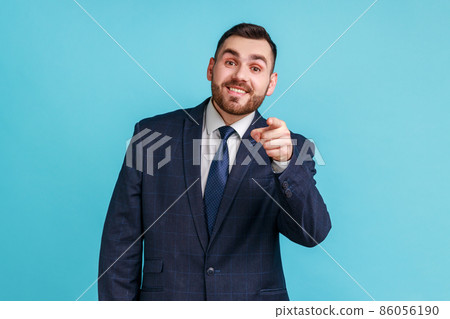 You are special. Young adult man with beard wearing official style suit pointing fingers at camera and looking with toothy smile, believes in you. Indoor studio shot isolated on blue background. 86056190