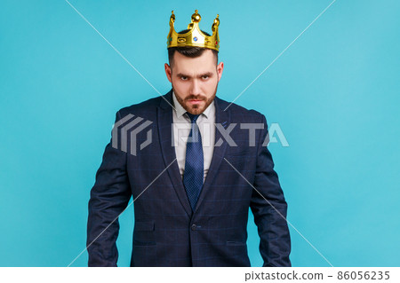 Strict bossy man in official style suit and golden crown, being on position of top manager or boss, looking with arrogance and anger, privileged status. Indoor studio shot isolated on blue background. 86056235