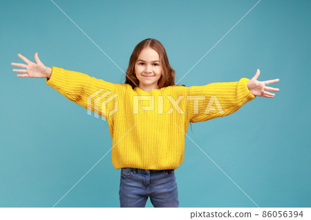 Portrait of happy cute little girl outstretching hands to embrace, giving free hugs and welcoming, wearing yellow casual style sweater. Indoor studio shot isolated on blue background. Portrait of happy cute little girl outstretching hands to embrace, giving free hugs and welcoming, wearing yellow casual style sweater. Indoor studio shot isolated on blue background. 86056394