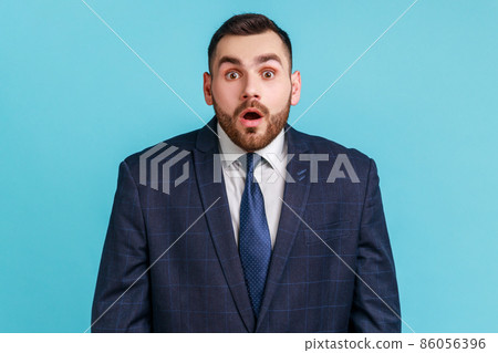 Wondered business man wearing official style suit looking at camera with shocked face and big eyes and open mouth, hearing astonishing news. Indoor studio shot isolated on blue background. Wondered business man wearing official style suit looking at camera with shocked face and big eyes and open mouth, hearing astonishing news. Indoor studio shot isolated on blue background. 86056396