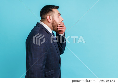 I won't say anyone. Side view of bearded man wearing official style suit covering mouth with hand, keeping terrible secret truth, don't want to talk. Indoor studio shot isolated on blue background. I won't say anyone. Side view of bearded man wearing official style suit covering mouth with hand, keeping terrible secret truth, don't want to talk. Indoor studio shot isolated on blue background. 86056423
