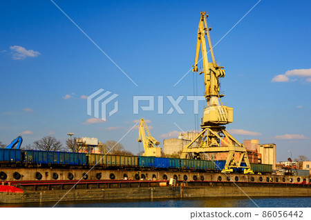 River port crane loading open-top gondola cars on sunny day. Empty river drag boats or barges moored by pier, Empty cars ready for loading. River port crane loading open-top gondola cars on sunny day. Empty river drag boats or barges moored by pier, Empty cars ready for loading. 86056442