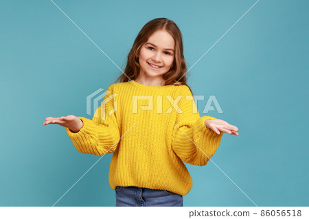 Portrait of little girl opens hands wide to give free hugs, ready to embrace friend, hospitable kind, wearing yellow casual style sweater. Indoor studio shot isolated on blue background. 86056518