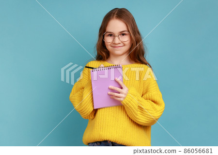 Portrait of positive little girl writing in notebook, looks smiling at camera, child doing homework, wearing yellow casual style sweater. Indoor studio shot isolated on blue background. 86056681