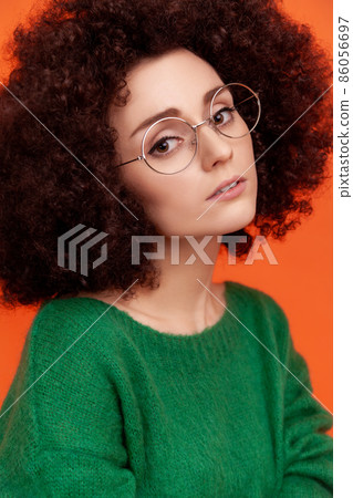 Portrait of pensive woman with perfect skin with Afro hairstyle wearing green casual style sweater, looking at camera, beauty and care concept. Indoor studio shot isolated on orange background. 86056697