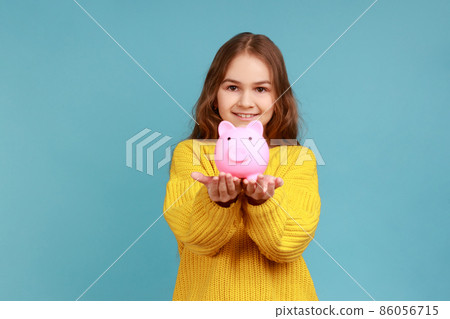 Portrait of smiling cute little girl holding out piggy bank to camera, happy with money savings, wearing yellow casual style sweater. Indoor studio shot isolated on blue background. 86056715