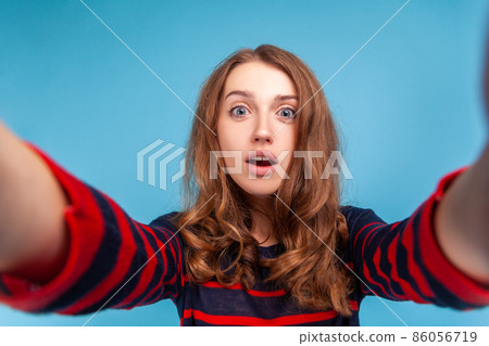 Woman with surprised facial expression taking selfie, point of view of photo, posing with open mouth, wearing striped casual style sweater. Indoor studio shot isolated on blue background. 86056719