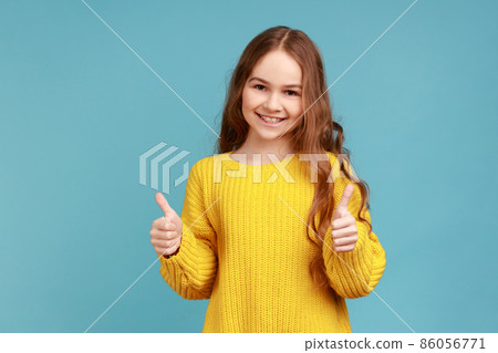 Portrait of charming little girl with wavy hair showing thumbs up to camera, expressing positive, wearing yellow casual style sweater. Indoor studio shot isolated on blue background. Portrait of charming little girl with wavy hair showing thumbs up to camera, expressing positive, wearing yellow casual style sweater. Indoor studio shot isolated on blue background. 86056771