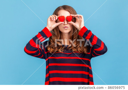 Romantic woman wearing striped casual style sweater, standing with pout lips, sending air kisses, covering eyes with small red heart love symbols. Indoor studio shot isolated on blue background. 86056975