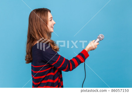 Side view portrait of woman wearing striped casual style sweater, standing with microphone in hands, offering for somebody to answer her questions. Indoor studio shot isolated on blue background. 86057150