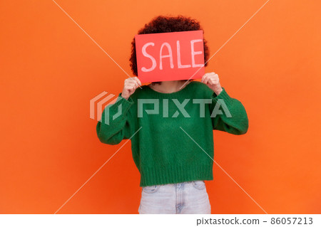 Portrait of unknown woman wearing green casual style sweater covering her face with card with sale inscriptions, shopping, discounts. Indoor studio shot isolated on orange background. Portrait of unknown woman wearing green casual style sweater covering her face with card with sale inscriptions, shopping, discounts. Indoor studio shot isolated on orange background. 86057213