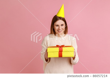 Portrait of blond woman in party cone holding yellow wrapped gift box, looking at camera with satisfied expression, wearing white sweater. Indoor studio shot isolated on pink background. Portrait of blond woman in party cone holding yellow wrapped gift box, looking at camera with satisfied expression, wearing white sweater. Indoor studio shot isolated on pink background. 86057214