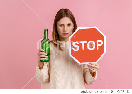 Portrait of serious blond woman holding red stop sign and bottle with alcoholic beverage, calls on dont drink alcohol, wearing white sweater. Indoor studio shot isolated on pink background. 86057238