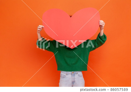 Unknown woman wearing green casual style sweater holding big red paper heart, hiding behind love huge love symbol, shy to express her feelings. Indoor studio shot isolated on orange background. 86057589