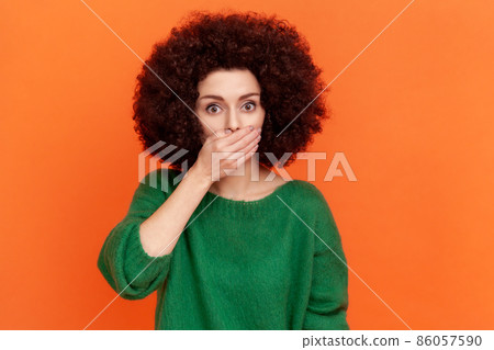 Young adult woman with Afro hairstyle wearing green casual style sweater covering mouth with palm, keep secret, does not tell anyone. Indoor studio shot isolated on orange background. Young adult woman with Afro hairstyle wearing green casual style sweater covering mouth with palm, keep secret, does not tell anyone. Indoor studio shot isolated on orange background. 86057590