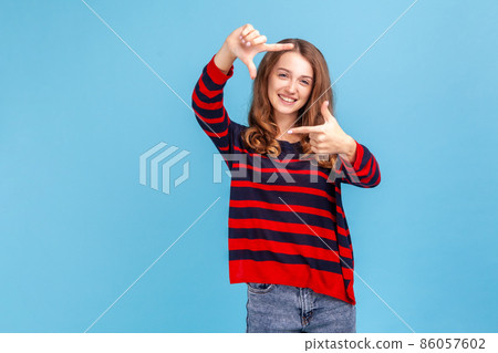 Smiling woman wearing striped casual style sweater looking at camera through hand frame, capturing photo or focusing eyesight on target. Indoor studio shot isolated on blue background. Smiling woman wearing striped casual style sweater looking at camera through hand frame, capturing photo or focusing eyesight on target. Indoor studio shot isolated on blue background. 86057602
