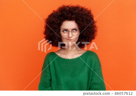 Portrait of confused woman with Afro hairstyle wearing green casual style sweater and optical glasses, looking away, frowning face, thinking. Indoor studio shot isolated on orange background. Portrait of confused woman with Afro hairstyle wearing green casual style sweater and optical glasses, looking away, frowning face, thinking. Indoor studio shot isolated on orange background. 86057612