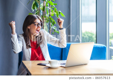 Extremely happy woman in glasses raising hands and screaming for joy, celebrating successful business online, rejoicing victory while working on laptop. Indoor shot, cafe or office background. Extremely happy woman in glasses raising hands and screaming for joy, celebrating successful business online, rejoicing victory while working on laptop. Indoor shot, cafe or office background. 86057613