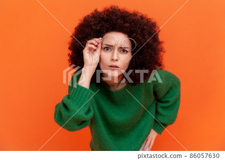 Curious woman with Afro hairstyle wearing green casual style sweater raising her optical glasses, wants to see something better, staring attentively. Indoor studio shot isolated on orange background. Curious woman with Afro hairstyle wearing green casual style sweater raising her optical glasses, wants to see something better, staring attentively. Indoor studio shot isolated on orange background. 86057630