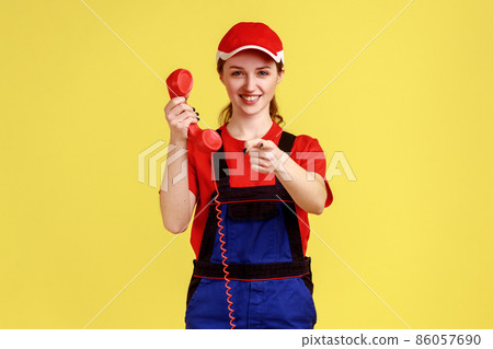 Portrait of smiling positive worker woman standing with landline phone in hands, pointing finger to camera, wearing overalls and red cap. Indoor studio shot isolated on yellow background. Portrait of smiling positive worker woman standing with landline phone in hands, pointing finger to camera, wearing overalls and red cap. Indoor studio shot isolated on yellow background. 86057690