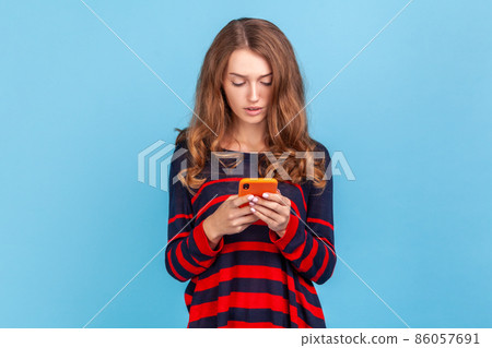 Portrait of serious woman wearing striped sweater, using smart phone, typing message or checking social networks, writing post, concentrated expression. Indoor studio shot isolated on blue background. Portrait of serious woman wearing striped sweater, using smart phone, typing message or checking social networks, writing post, concentrated expression. Indoor studio shot isolated on blue background. 86057691