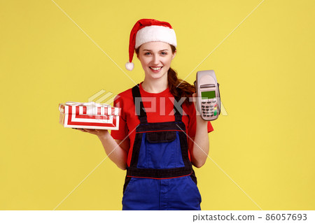 Optimistic delivery woman showing gift box and terminal, contactless payment for online ordered goods, wearing blue overalls and santa claus hat. Indoor studio shot isolated on yellow background. Optimistic delivery woman showing gift box and terminal, contactless payment for online ordered goods, wearing blue overalls and santa claus hat. Indoor studio shot isolated on yellow background. 86057693