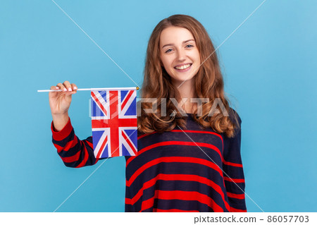 Attractive woman wearing striped casual style sweater, holding flag of a constituent unit of the United Kingdom, celebrating British Independence Day Indoor studio shot isolated on blue background. Attractive woman wearing striped casual style sweater, holding flag of a constituent unit of the United Kingdom, celebrating British Independence Day Indoor studio shot isolated on blue background. 86057703