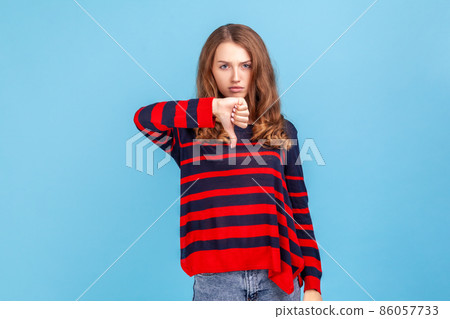 Unhappy unsatisfied young woman wearing striped casual style sweater showing thumbs down, dislike gesture, negative emotions, bad rating. Indoor studio shot isolated on blue background. 86057733