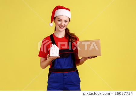 Smiling woman mover or worker holding cardboard parcel and paper house, relocation service, wearing blue overalls and santa claus hat. Indoor studio shot isolated on yellow background. Smiling woman mover or worker holding cardboard parcel and paper house, relocation service, wearing blue overalls and santa claus hat. Indoor studio shot isolated on yellow background. 86057734