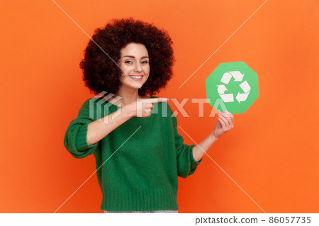 Satisfied woman with Afro hairstyle wearing green casual style sweater pointing at green recycling symbol in her hands, ecology concept. Indoor studio shot isolated on orange background. 86057735