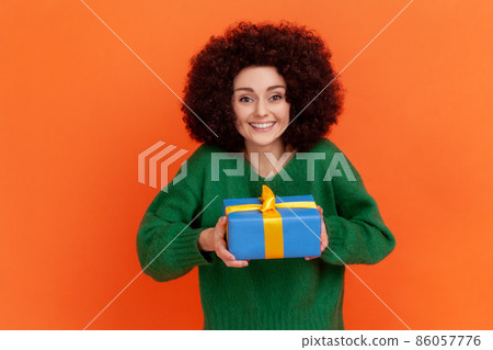 Happy positive smiling woman with Afro hairstyle wearing green casual style sweater holding wrapped gift box, looking at camera, congratulating. Indoor studio shot isolated on orange background. Happy positive smiling woman with Afro hairstyle wearing green casual style sweater holding wrapped gift box, looking at camera, congratulating. Indoor studio shot isolated on orange background. 86057776