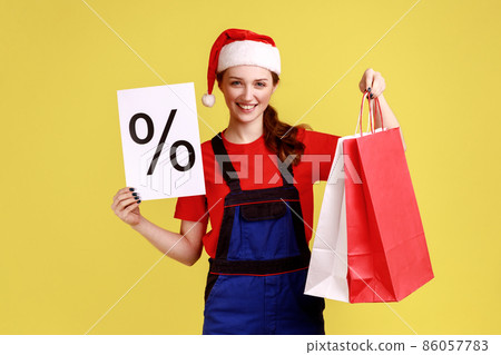 Positive courier female shows paper with percent mark, shows shopping bags, discounts for delivering, wearing blue overalls and santa claus hat. Indoor studio shot isolated on yellow background. Positive courier female shows paper with percent mark, shows shopping bags, discounts for delivering, wearing blue overalls and santa claus hat. Indoor studio shot isolated on yellow background. 86057783