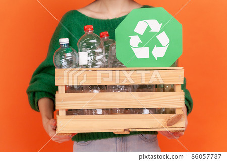 Anonymous woman wearing green casual style sweater holding box with plastic bottles ans green recycling symbol, ecological problems. Indoor studio shot isolated on orange background. 86057787