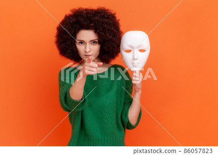 Strict bossy woman with Afro hairstyle in green casual style sweater holding white mask in hand, pointing finger to camera with serious expression. Indoor studio shot isolated on orange background. 86057823