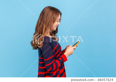Side view of smiling optimistic woman standing with smart phone in hands, answering romantic message from boyfriend, wearing striped casual sweater, Indoor studio shot isolated on blue background 86057824