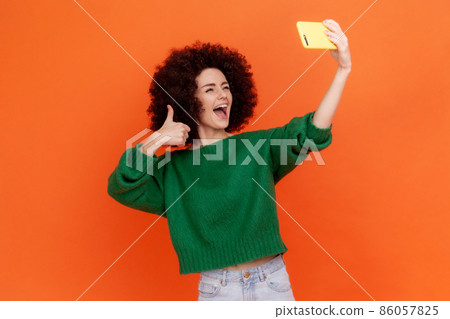 Excited happy woman with Afro hairstyle wearing green casual style sweater has livestream with followers, showing thumb up to camera. Indoor studio shot isolated on orange background. 86057825