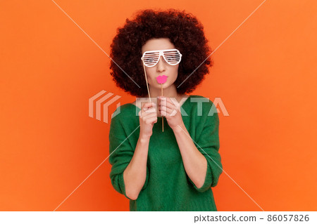 Funny woman with Afro hairstyle wearing green casual style sweater covering eyes with paper glasses, holding red lips on stick, masquerade. Indoor studio shot isolated on orange background. Funny woman with Afro hairstyle wearing green casual style sweater covering eyes with paper glasses, holding red lips on stick, masquerade. Indoor studio shot isolated on orange background. 86057826