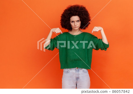 Here and right now. Serious woman with Afro hairstyle wearing green casual style sweater standing and looking at camera, pointing down. Indoor studio shot isolated on orange background. 86057842