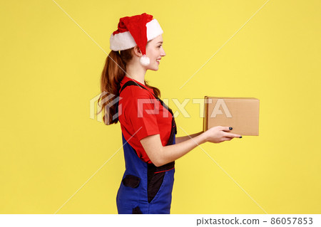 Side view portrait of courier female handing cardboard parcel for client, present for New Year, wearing blue overalls and santa claus hat. Indoor studio shot isolated on yellow background. 86057853