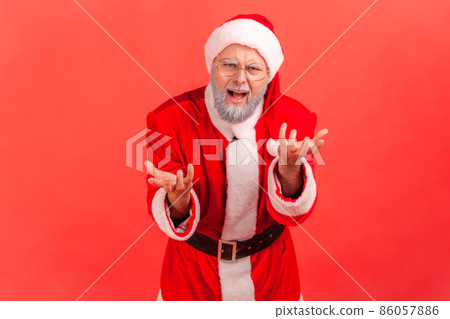 Portrait of elderly man with gray beard wearing santa claus costume standing with raised hands and indignant aggressive face asking why. Indoor studio shot isolated on red background. 86057886