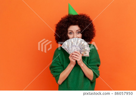 Happy woman with Afro hairstyle wearing green casual style sweater holding covering half of mouth with money, having big sum of money as birthday gift. Indoor studio shot isolated on orange background 86057950