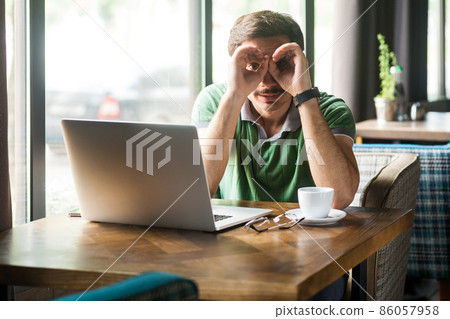Portrait of businessman wearing green T-shirt, working online on laptop, looking through fingers shaped like binoculars and expressing positive. Indoor shot near big window, cafe background. Portrait of businessman wearing green T-shirt, working online on laptop, looking through fingers shaped like binoculars and expressing positive. Indoor shot near big window, cafe background. 86057958