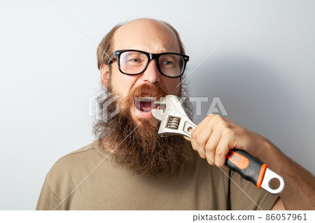 Portrait of male trying to pull out bad tooth with adjustable wrench , keeps mouth opened, bald bearded man wearing T-shirt and glasses. Indoor studio shot isolated on gray background. 86057961
