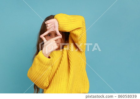 Portrait of little girl looking through photo frame hand gesture, focusing picture, observing world, wearing yellow casual style sweater. Indoor studio shot isolated on blue background. Portrait of little girl looking through photo frame hand gesture, focusing picture, observing world, wearing yellow casual style sweater. Indoor studio shot isolated on blue background. 86058039