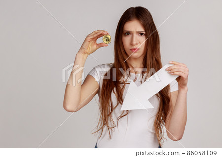 Portrait of unhappy woman holding golden bitcoin and white paper arrow, points down, showing downgrade of cryptocurrency, wearing white T-shirt. Indoor studio shot isolated on gray background. 86058109